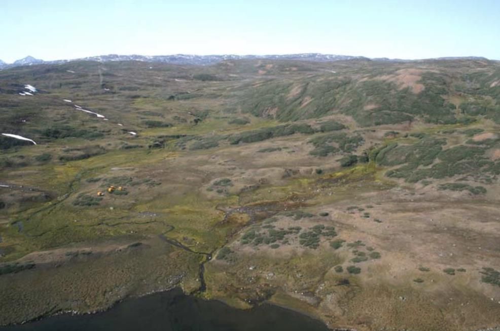 Aerial view over the Qeqertaasaq project area: low green hills and tundra beside a lake, with an exploration camp in the middle distance